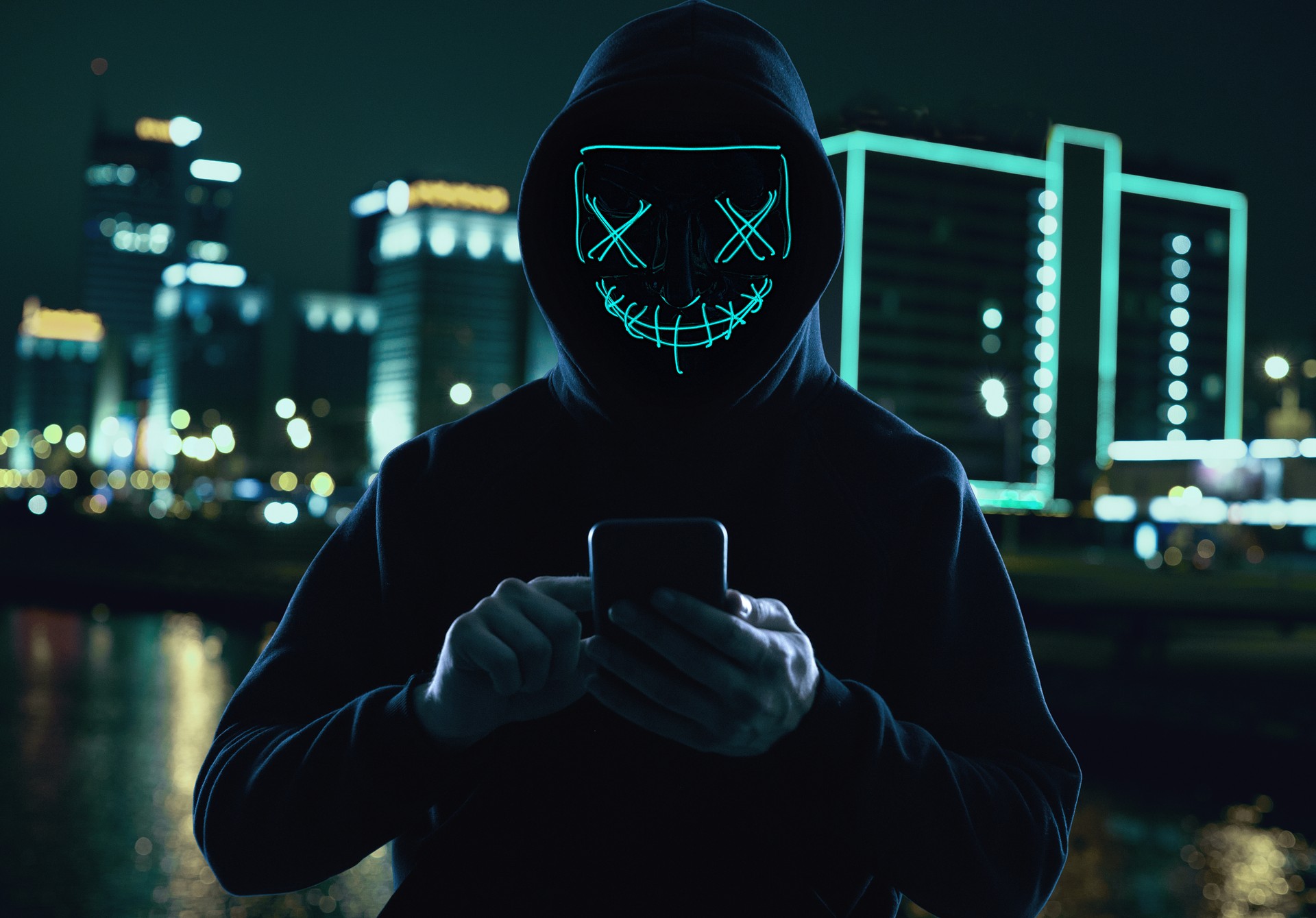 Anonymous man in a black hoodie and neon mask hacking into a smartphone