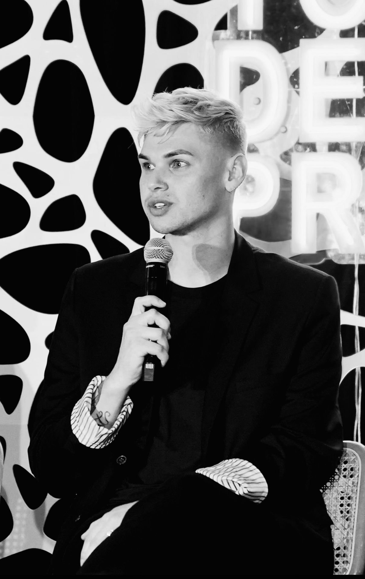 Person dressed in black holding a microphone while seated against a patterned backdrop and neon sign.
